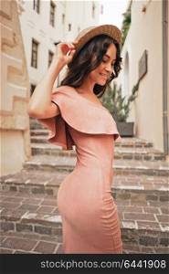 Beautiful brunette young woman wearing pink dress and straw hat walking on the street in old european Town. Fashion and style. Summer travel