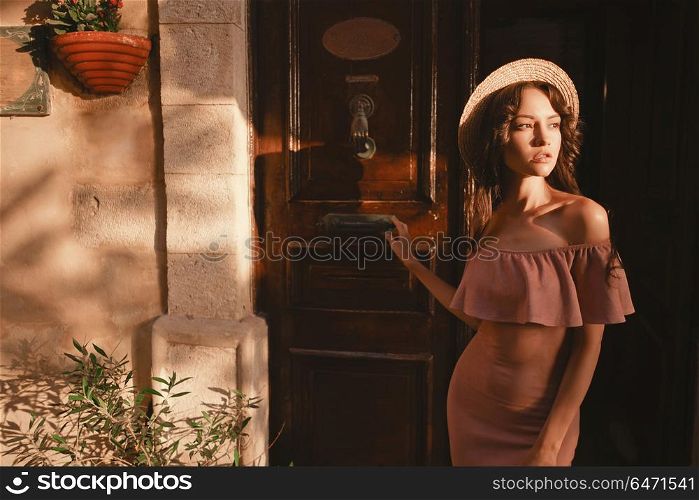 Beautiful brunette young woman wearing pink dress and straw hat walking on the street in old european Town. Fashion and style. Summer travel. Stylish brunette woman walking in old town