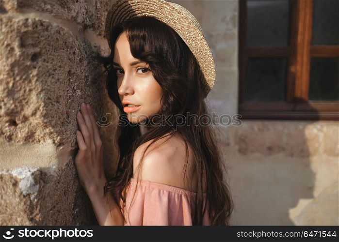 Beautiful brunette young woman wearing pink dress and straw hat walking on the street in old european Town. Fashion and style. Summer travel. Stylish brunette woman walking in old town