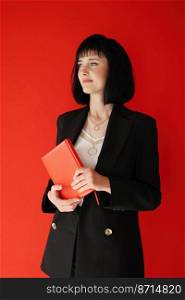 Beautiful business woman holding paper notebook isolated on red background. Successful brunette female in black suit with short hair is looking in camera. Business, career success, education concept. Beautiful business woman holding paper notebook isolated on red background. Successful brunette female in black suit with short hair is looking in camera. Business, career success, education concept.