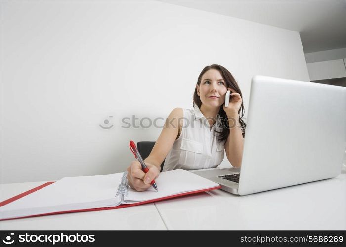 Beautiful businesswoman answering cell phone while writing at desk in office