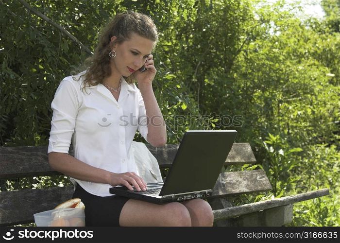 Beautiful businesswoman having her lunchbreak in the park while working on her laptop and talking on the phone