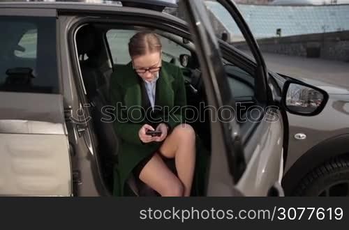 Beautiful businesswoman sending message on smart phone while sitting in car seat with opened door in daylight. Elegant blond woman with perfect legs in high heels shoes wearing emerald green coat and eyeglasses using mobile phone in auto. Slow motion