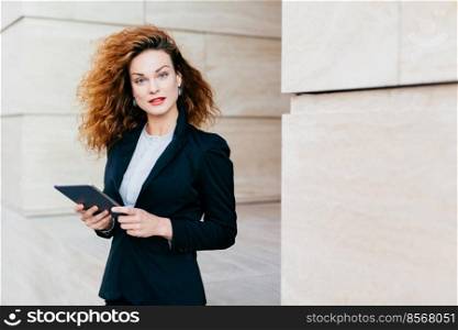 Beautiful businesswoman with curly fluffy hair, wearing elegant clothes, holding tablet in hands, waiting for her business partner near cafe, using free internet conncection. People, career, work