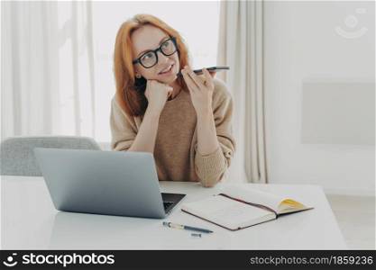Beautiful busy ginger female freelancer records voice message holds mobile phone near mouth sits at desktop makes notes in notepad works on laptop computer. People technology and job concept. female freelancer records voice message sits at desktop makes notes in notepad