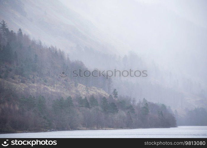 Beautiful calm peaceful Winter landscape over Thirlmere in Lake District with fog and layers of trees visible in the distance