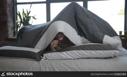 Beautiful carefree family with cute daughter looking out from under the blanket and smiling. Positive little girl with parents posing under devut in modern bedroom in the morning, Joyful family having fun and playing together. Slo mo. Dolly shot.