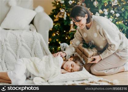 Beautiful caring mother prepares present box for little child who sleeps on floor near Christmas tree, makes surprise for holiday, stands on knees near daughter. Motherhood, children, New Year time