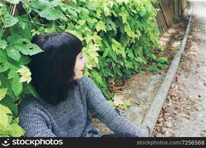 Beautiful caucasian brunette woman sitting on ground in a park wearing punk or gothic clothes and surrounded by green leafs