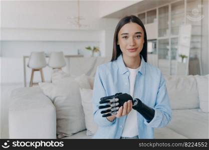 Beautiful caucasian girl with disability taking apart her bionic prosthetic arm, standing at home. Smiling disabled young woman showing high tech robotic hand, looking at camera in living room.. Beautiful caucasian girl with disability taking apart her bionic prosthetic arm, standing at home.