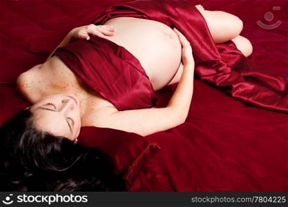 Beautiful Caucasian pregnant brunette woman holding her bare belly laying in a bed with red satin sheets during pregnancy.