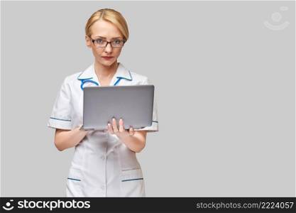 Beautiful Caucasian woman doctor or nurse holding a laptop pc computer standing over grey background.. Beautiful Caucasian woman doctor or nurse holding a laptop pc computer standing over grey background