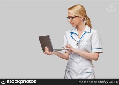Beautiful Caucasian woman doctor or nurse holding a laptop pc computer standing over grey background.. Beautiful Caucasian woman doctor or nurse holding a laptop pc computer standing over grey background