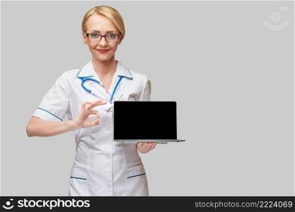Beautiful Caucasian woman doctor or nurse holding a laptop pc computer with blank screen standing over grey background.. Beautiful Caucasian woman doctor or nurse holding a laptop pc computer with blank screen standing over grey background