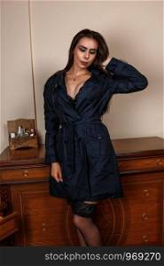 beautiful Caucasian woman housewife wife in black raincoat and in beautiful lingerie stands next to the dressing table at home