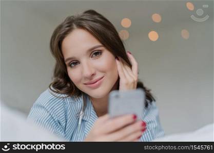 Beautiful Caucasian woman listens audio with earphones, connected to modern smart phone device, watches video in social networks, lies in bed indoor. People, technology and entertainment concept