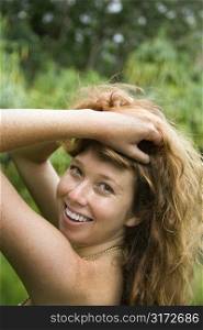 Beautiful Caucasian young adult woman in lush forest holding up long wavy hair smiling and looking at viewer.