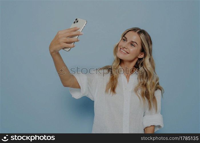 Beautiful caucasian young female with long blonde dyed hair holding mobile phone and posing for selfie, looking at screen with flirty smile, attractive woman making photo for social networks online. Beautiful young female with long blonde dyed hair holding mobile phone and posing for selfie