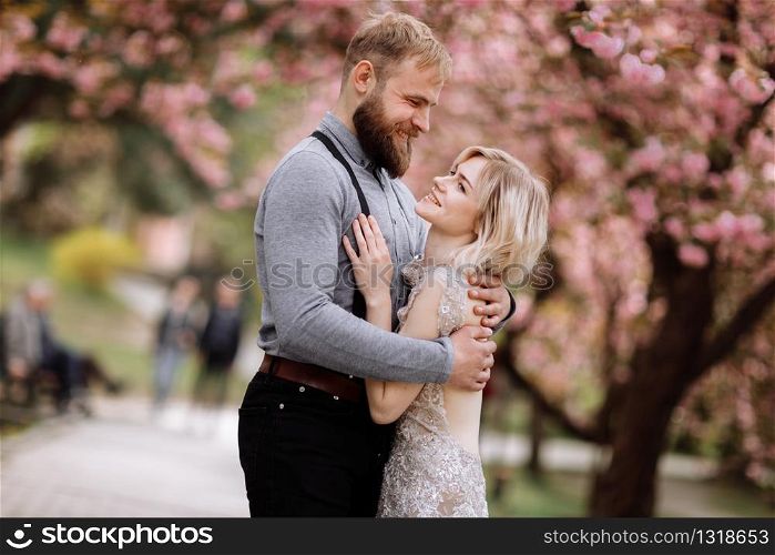 Beautiful, cheerful and cute couple in blossoming pink cherry blossom, sakura garden, hugging and looking to each other on a sunny day. Spring wedding portrait.. Beautiful, cheerful and cute couple in blossoming pink cherry blossom, sakura garden, hugging and looking to each other on a sunny day. Spring wedding portrait