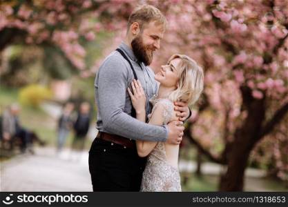 Beautiful, cheerful and cute couple in blossoming pink cherry blossom, sakura garden, hugging and looking to each other on a sunny day. Spring wedding portrait.. Beautiful, cheerful and cute couple in blossoming pink cherry blossom, sakura garden, hugging and looking to each other on a sunny day. Spring wedding portrait