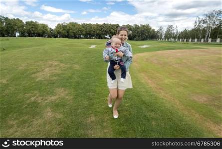 Beautiful cheerful mother holding her baby boy and running on grass at park