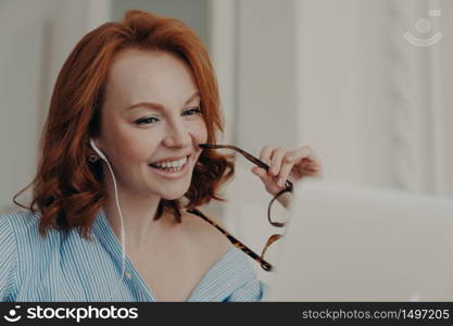 Beautiful cheerful redhead woman uses laptop computer for online conference, talks with colleagues distantly, holds spectacles, discusses working issues, works freelance from home. Technology concept