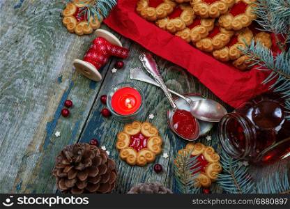Beautiful Christmas card: Sweet Christmas cookies with jam, fir branches and cones, cranberries, red burning candle and the bobbin with decorative ribbon on the old wooden table, with copy-space