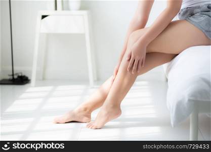 Beautiful closeup young asian woman sitting on a bed stroking legs with soft smooth skin in the bedroom, girl applying body cream and lotion with treatment, healthy and wellness concept.