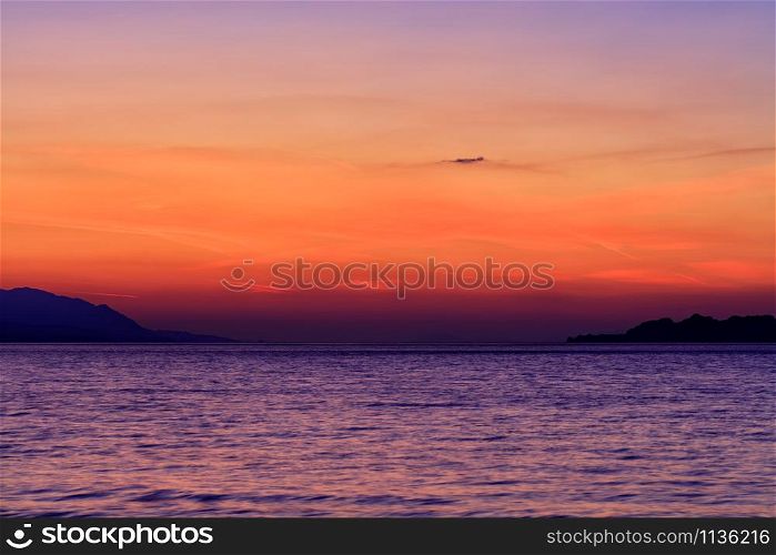 Beautiful, colorful sunset on the sea, mountains silhouettes and bright traces of tracks of flying planes in the sky, image with copy space.. Beautiful, bright sunset on the Gulf of Corinth against the background of the silhouette of the highlands running along the horizon of the sea line.