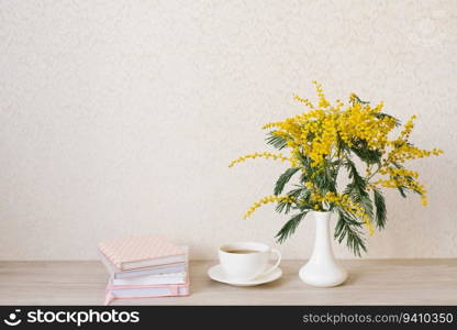 Beautiful composition of mimosa branches and a white tea cup with a saucer and a stack of notebooks on a table with a copy space