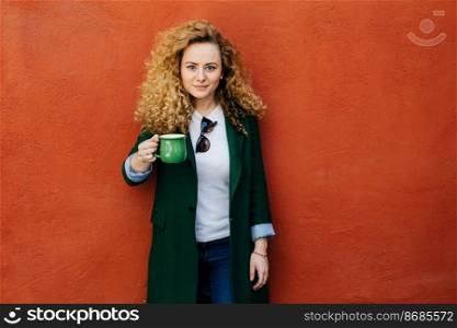 Beautiful confident blonde curly woman wearing elegant jacket and jeans holding green mug with tea posing against orange background in studio. People, free time, lifestyle and relaxation concept