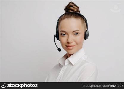 Beautiful consultant in white shirt, black headset, hair in bun, discusses client&rsquo;s insurance fund online, isolated near light wall.