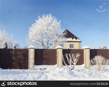 Beautiful country landscape on frosty winter day. Stone cottage behind the metal fence, bare trees covered with hoarfrost.