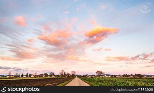 Beautiful countryside landscape with road, fields and blue sky covered partially with fluffy colorful clouds in the background.