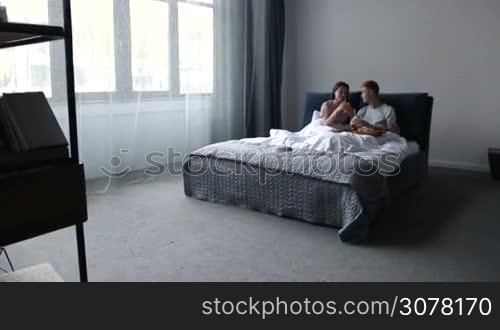 Beautiful couple in love lying in bed and drinking orange juice with straws from mason jars during romantic breakfast in the morning. Smiling couple enjoying leisure in bedroom, talking and having breakfast served on wooden tray. Slow motion.