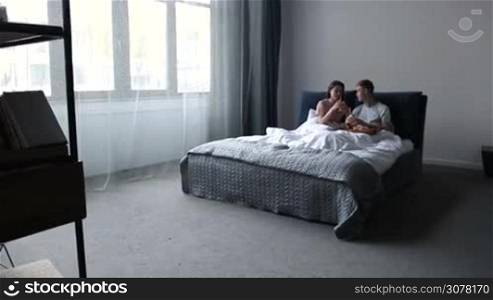 Beautiful couple in love lying in bed and drinking orange juice with straws from mason jars during romantic breakfast in the morning. Smiling couple enjoying leisure in bedroom, talking and having breakfast served on wooden tray. Slow motion.