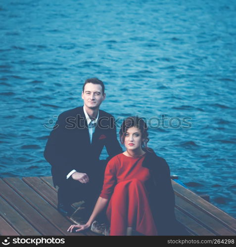 Beautiful couple of lovers - a man in a suit and a woman in a red dress sitting on a pier against a ripple of water. Vintage blue toned filter, square portrait with space for copy.. Blue Toned Portrait Of A Couple