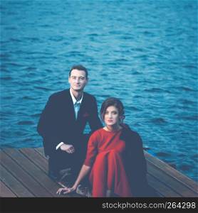 Beautiful couple of lovers - a man in a suit and a woman in a red dress sitting on a pier against a ripple of water. Vintage blue toned filter, square portrait with space for copy.. Blue Toned Portrait Of A Couple