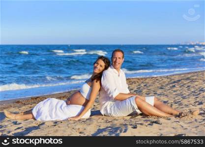 Beautiful couple pregnant woman in the beach sitting on sand happy together back to back