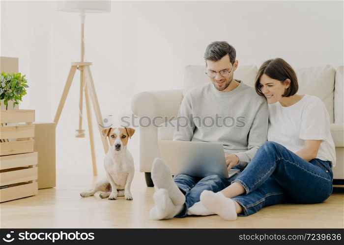 Beautiful couple with dog sit on floor in spacious living room, make purchasing online, buy furniture for new home, have to unpack belongings, have happy expression, start living together in new flat