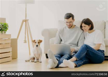 Beautiful couple with dog sit on floor in spacious living room, make purchasing online, buy furniture for new home, have to unpack belongings, have happy expression, start living together in new flat