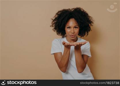 Beautiful curly flirty african woman in white t-shirt sending air kiss and smiling, demonstrating warm feelings and love while posing in studio on beige background with copy space for text. Beautiful curly flirty african woman sending air kiss and smiling while posing on beige background