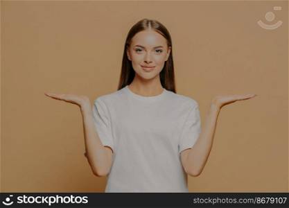 Beautiful dark haired European woman raises palms pretends holding something over blank copy space against brown background wears casual white t shirt proposes product. Advertisement gesture