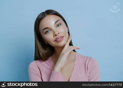 Beautiful dark haired woman with applied makeup, healthy glowing skin, touches cheek gently, wears rosy jumper, looks directly at camera, isolated over blue background. Women and beauty concept