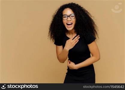 Beautiful dark skinned model cannot stop laughing, hears comic story, keeps hand on belly, laughs loudly, dressed in black clothes, stands against brown studio wall with empty space for information