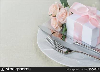 Beautiful decorated table with white plates, gift box with a pink ribbon, cutlery and pink rose flowers on tablecloths, with space for text