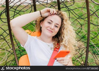 Beautiful delighted female blogger with curly light hair, uses smart phone for posting new posts on her web site, rests in hanging chair, uses wireless internet connection. People, recreation concept