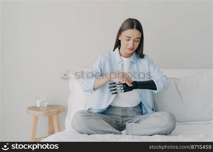 Beautiful disabled caucasian girl setting her bionic arm prosthesis, sitting on bed at home. Modern young woman touching artificial robotic hand in bedroom. Lifestyle of people with disabilities.. Disabled girl setting bionic arm prosthesis, sitting on bed. Lifestyle of people with disabilities