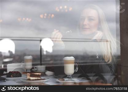 Beautiful dreamy woman sitting in cafe and with pleasure eating dessert and drinking latte, enjoying time in the restaurant, modern life of young people