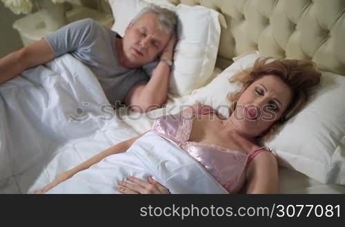 Beautiful elderly woman stretching and smiling while waking up in her bed at home. Charming senior woman with her head resting on the white pillow relaxing on bed and looking at camera while her husband is still sleeping peacefully.
